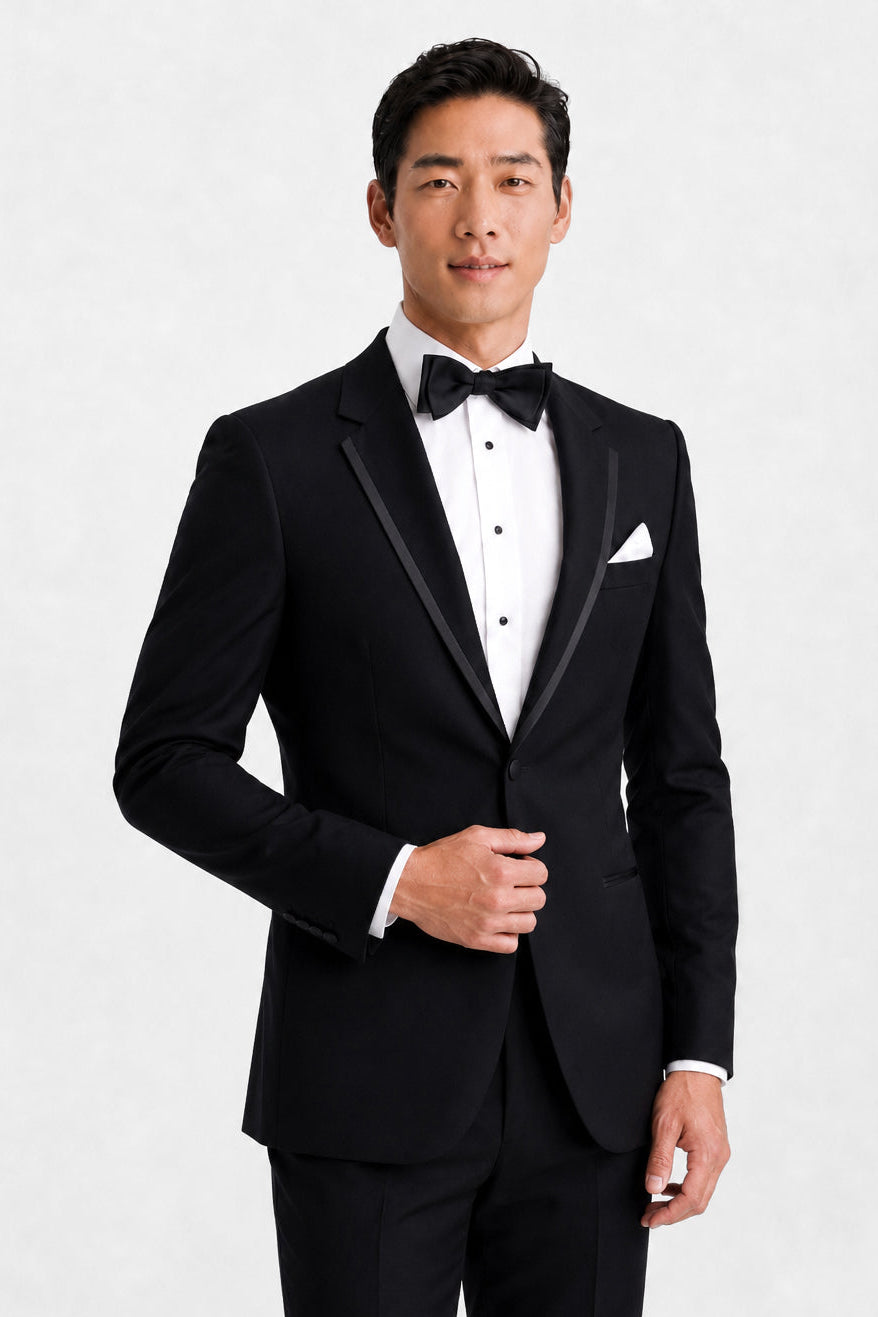 A man modeling a classic black tuxedo with satin lapels, a white dress shirt, a black bow tie, and a white pocket square, shot against a light gray studio background.