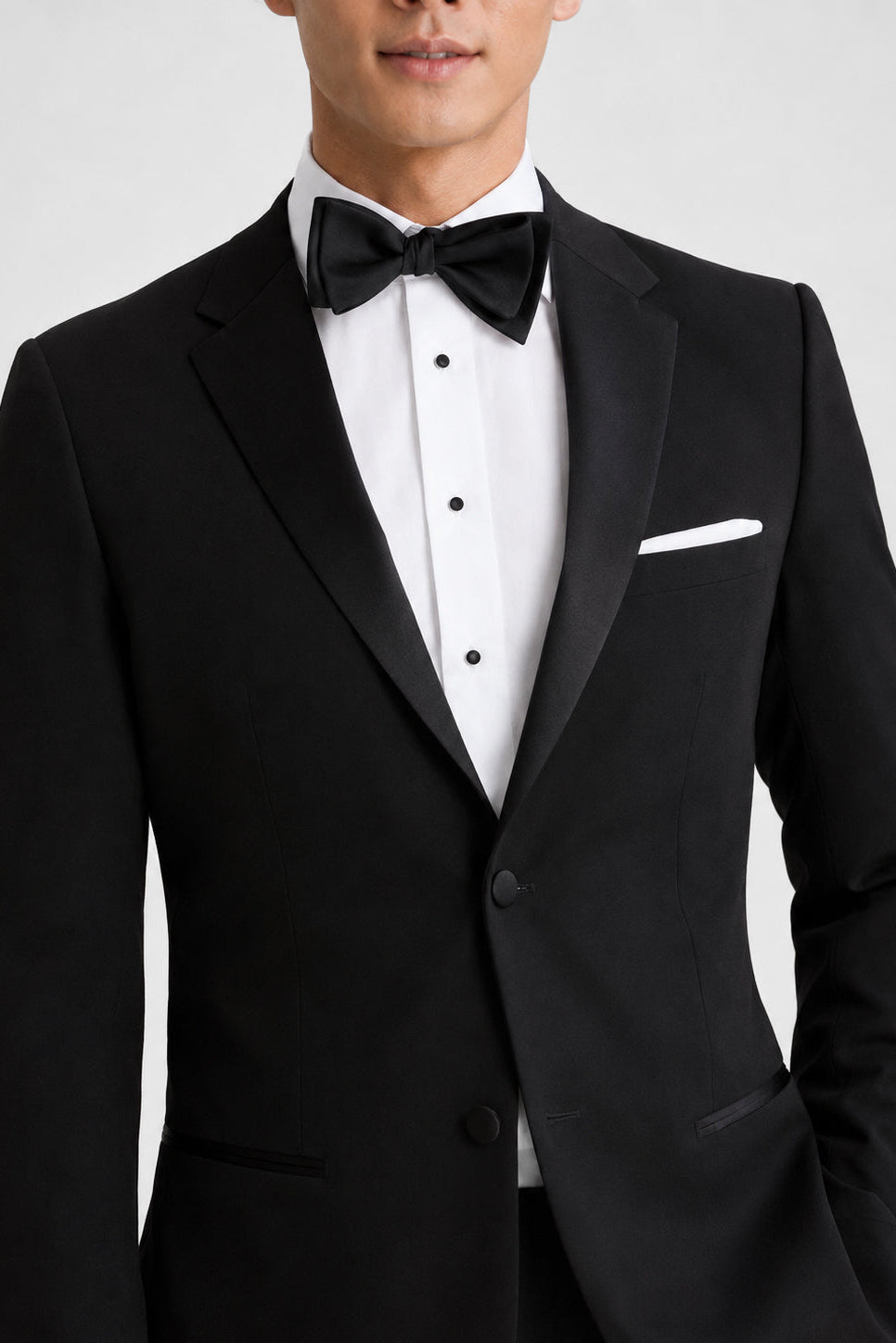A man wearing a black tuxedo jacket with satin lapels, a white dress shirt, a black bow tie, and a white pocket square, shown against a light gray studio background.