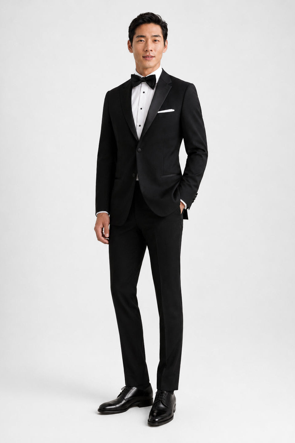 A man modeling a classic black tuxedo with satin lapels, a white dress shirt, a black bow tie, and a white pocket square, set against a plain light gray studio background.