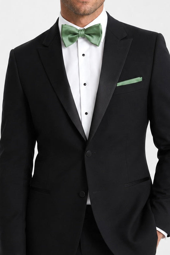 A man wearing a black tuxedo jacket with satin lapels, a white tuxedo shirt, a green bow tie, and a matching green pocket square, photographed against a light gray studio background.