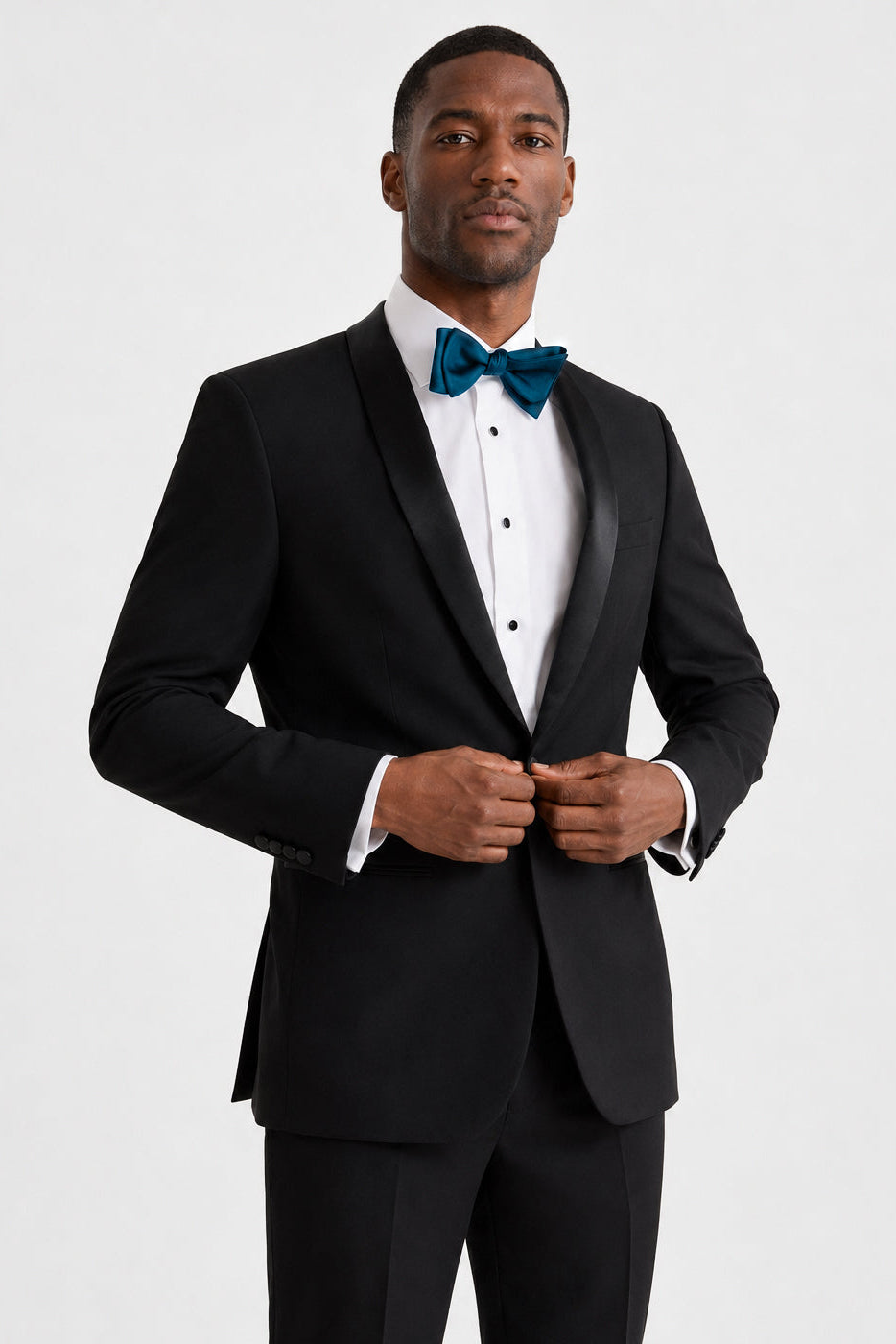 A man wearing a black tuxedo jacket with satin peak lapels, a white pleated tuxedo shirt, and a teal bow tie, standing against a light gray studio background.